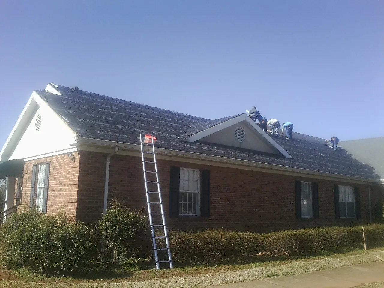 Crew tearing shingles off a brick ranch roof with a ladder set — Metal Roof Repair work by our crew in New Freeport