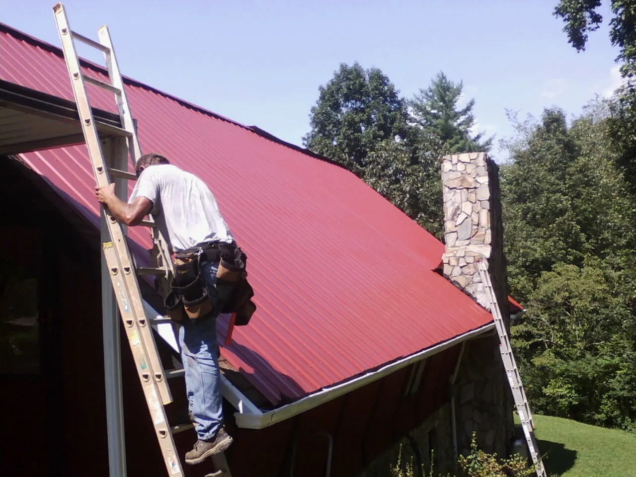 Roofer on a ladder beside a red metal roof and stone chimney — Soffit & Fascia Repair by Roofing Brothers in New Freeport